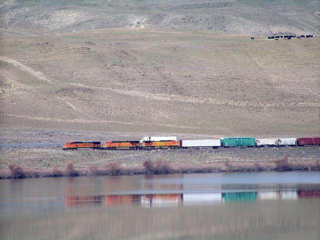 A train being chased by a semi across the river. The water is quiet enough to reflect the train.